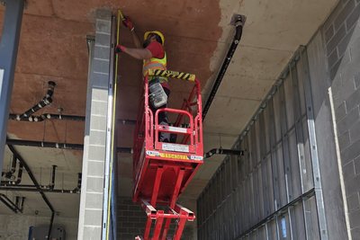 An installer in a high-visibility vest working from a scissor lift, measuring at the head of a steel stud wall under a concrete slab.