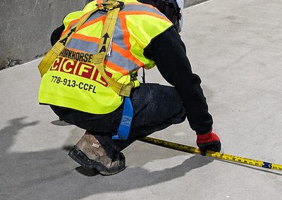 An installer in a high-visibility vest kneeling on bare concrete, pulling a tape measure along a wall layout.